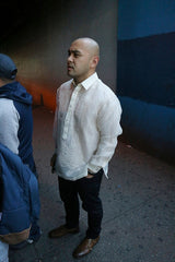 Steven wears a hand embroidered piña silk Barong Tagalog, a chamisa de chino underneath his barong, dark jeans and brown dress shoes. He stands with his hands in his pockets on the sidewalk behind a couple people on a line. There is a concrete wall, part of which is painted blue, behind him.