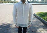 Product photo of the Steven barong. Steven stands in Flushing Meadow Park in front of the base of the Unisphere. Steven wears a hand embroidered piña silk Barong Tagalog, a chamisa de chino underneath his barong, and dark jeans. He stands on a paving stone walkway next to the grass and there are trees, the base of the Unisphere and the blue sky in the background. Steven's face is kept out of the picture. His hands are hanging just outside his pockets
