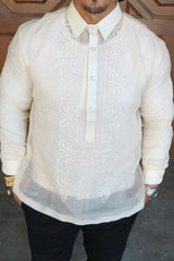 Product photo of the hand embroidered piña silk Zar Barong Tagalog. Zar wears a chamisa de chino underneath his barong, beaded bracelet on right wrist and watch on left wrist, and black pants. Zar stands in front of brown wooden doors