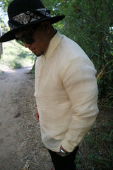 Picture of side view of Zar standing with head tilted down with hands in pockets on a dirt path with trees and branches in the background. Zar wears a hand embroidered piña silk Barong Tagalog, chamisa de chino underneath his barong, black sunglasses and black hat with black bandana tied around the hat, watch on left wrist, and black pants.