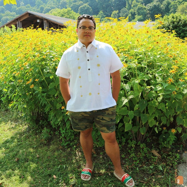 Chef Dale Talde stands in front of many yellow flowers in his white ramie everyday Barong Tagalog with sun and stars necklace gold embroidery. He also wears camo shorts and Gucci slides.