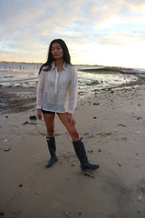 Andrelie standing in her custom hand embroidered jusi Barong Tagalog, black shorts and black boots on the sand with her hand on her leg and the water behind her at Dead Horse Bay Beach in Brooklyn, NY