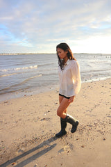 Andrelie walking in her custom hand embroidered jusi Barong Tagalog, black shorts and black boots on the sand next to the water at Dead Horse Bay Beach in Brooklyn, NY