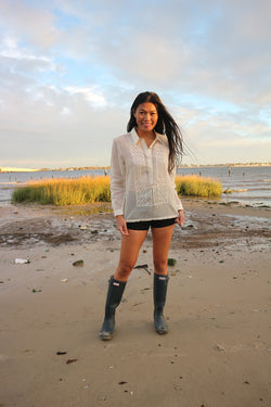 Andrelie standing in her custom hand embroidered jusi Barong Tagalog, black shorts and black boots on the sand with tall grass and water behind her at Dead Horse Bay Beach in Brooklyn, NY