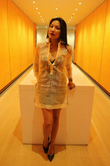 Anna stands and leans with hands resting on white table behind her in orange hallway. Anna wears a dress length hand embroidered piña silk Barong Tagalog 