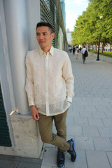 Arland stands and leans on grey pillar in his long sleeve full button down hand embroidered piña silk Barong Tagalog, olive green pants and black shoes. There are people walking and trees behind Arland in the distance. Arland is at the Jersey City Waterfront