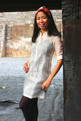 Bianca stands, smiles and leans on dark wood pillar in her hand embroidered cocoon Barong Tagalog, with black leggings and red and white headband with a stone wall in the background