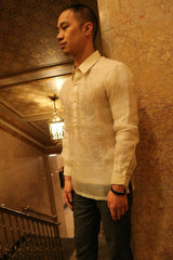 Side angled view of Chris M from the knees up standing with his back leaning on a marble wall next to a staircase, hand railing, patterned ceiling and light fixture. Chris M wears his hand embroidered piña silk Barong Tagalog, dark pants and black bracelet. Chris looks straight and down with his hands at his sides. 