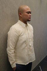 Side angle view of Chris U leaning his back to a grey patterned wall with Chris looking straight ahead with his hands in his pockets wearing a hand embroidered jusi Barong Tagalog and dark blue slacks