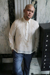 Chris U stands and leans against mailbox with a marble wall behind him with his hands in his pockets wearing a hand embroidered jusi Barong Tagalog and blue slacks.