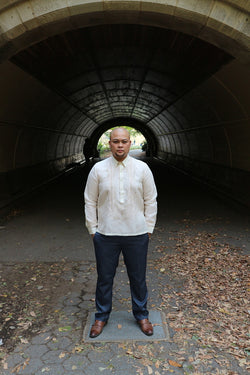 Chris U stands in front of a tunnel in Prospect Park, Brooklyn, with his hands in his pockets wearing his hand embroidered jusi Barong Tagalog, blue slacks and brown dress shoes