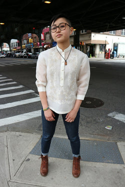 Chrissi stands at the corner of 69 Street and Roosevelt Avenue in Little Manila in Woodside, NY. Chrissi wears a hand embroidered jusi Barong Tagalog, blue jeans, brown boots, glasses and a necklace.