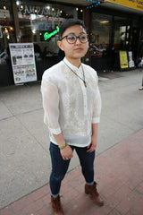 Angled shot of Chrissi wearing a hand embroidered jusi Barong Tagalog, blue jeans, brown boots, glasses and a necklace while standing on the sidewalk in front of Renee's restaurant in Little Manila in Woodside, NY