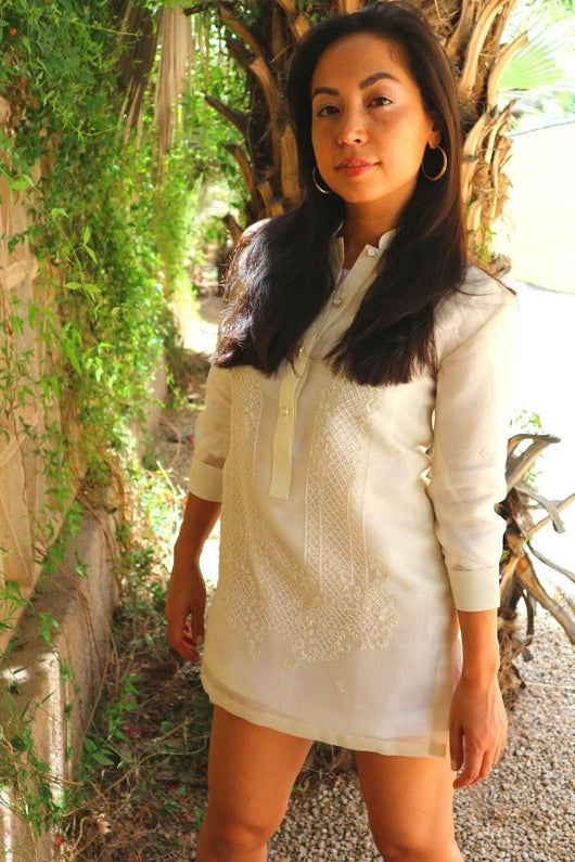 Coley from the knees up wearing a dress length hand embroidered cocoon Barong Tagalog next to a wall with green vines and leaves hanging off of it and a palm tree trunk behind her