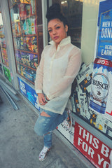 Dom leans up against a bodega window with beer advertising behind her. Dom wears a dress length hand embroidered cocoon barong Tagalog, ripped blue jeans, white and patterned Nike Cortez, rings, bracelets and earrings. Dom stands with her left leg pressed against the bottom of the window and her hands together covering the bottom center part of her barong. Dom's hair is in a bun on top of her head   