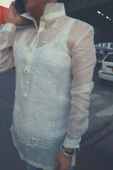 Angled photo of Dom on the phone but the shot focuses on the hand embroidered cocoon Barong Tagalog's front left side. Dom holds her cell phone to her ear with her right hand, her Barong's collar stands up, she wears bracelets on her left wrist 
