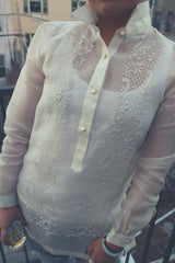 Product shot of the full front of the hand embroidered cocoon Dom Barong Tagalog. Dom stands at an angle with her hands at her sides holding a glass of whiskey in her right hand. Dom's collar on her barong is standing up.