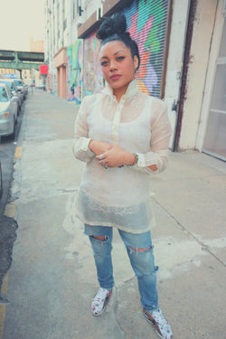 Dom stands on the sidewalk wearing a dress length hand embroidered cocoon Barong Tagalog with ripped blue jeans, multicolored Nike Cortez's, bracelets, rings and earrings with her hair in a bun on top of her head. Dom stands holding her hands in front of her with buildings and colorful murals behind her
