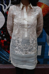 Product picture of the dress length Iris Z hand embroidered piña silk Barong Tagalog. Model has her hands behind her back and stands before a multi-colored mural