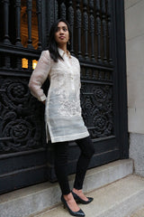 Angled full body picture of Iris Z standing with her back to black cast iron decorative gates. Iris stands with her hands behind her back and looking to her right. She wears a dress length hand embroidered piña silk Barong Tagalog, with beige camisole, black leggings and black high heeled shoes underneath