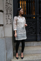 Front full body picture of Iris Z standing on stairs in front of a black cast iron decorative gate and a carved stone wall to her right. Iris stands with her left hand lifted next to her waist, right hand down at her side and looking to her left. She wears a dress length hand embroidered piña silk Barong Tagalog, with beige camisole, black leggings and black shoes underneath