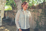 Iris B stands in front of a tree with wooden fence with green vines, and other trees behind them. Iris wears her blouse length full button down hand embroidered piña silk Barong Tagalog. She also wears black feather earrings, silver bracelets and black dress underneath her barong 
