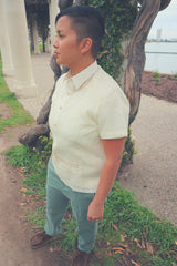 Photo of a side view of Irma standing on grass and soil next to a column covered by plants and a tree trunk. Irma wears a short sleeve embroidered jusi Barong Tagalog, green khaki pants and brown Clark Wallabee shoes. There is a walkway path, white columns, Lake Merritt and buildings in the background