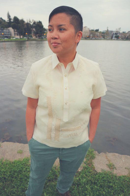 Irma stands in front of Lake Merritt with her hands in her pockets and she looks away. Irma wears a jusi short sleeve Barong Tagalog and green khaki pants. The lake and homes are in the background.