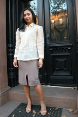 Jenny stands in front of black doors with windows reflecting the brick building across the street. Jenny wears a hand embroidered piña silk Barong Tagalog, grey skirt and pink open toe shoes