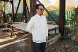 John-T stands leaning his back on a brown steel beam with a wooden table, more steel beams, green buildings and trees behind him. John-T wears a hand embroidered jusi Barong Tagalog, dark sunglasses, dark blue jeans. John-T stands at an angle with his right hand behind his back and left hand at his side. There are wooden planters and potted plants to his left
