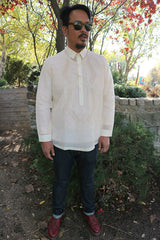 Full body picture of John-T standing on soil with shrubs trees, and walls of wood and brick in the background. John-T wears a hand embroidered jusi Barong Tagalog, dark sunglasses, dark blue jeans and dark red Doc Marten boots. There are red and light green leaves on the trees behind him