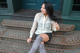 Kristina sits on brown stairs textured with circles in front of a green doorway. Kristina wears a hand embroidered piña silk Barong Tagalog, a black tank top underneath her barong, black perforated leather miniskirt, gold bracelets on each wrist and you can see the top half of her lavendar suede thigh high boots. Kristina has her legs crossed, she smiles and looks away. Her right hand rests on the stair and her left hand holds the ends of her hair