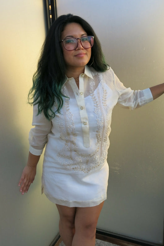 Krystal wearing a dress length hand embroidered jusi Barong Tagalog. She stands between 2 panels of frosted glass. She wears plastic rimmed glasses.