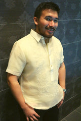 Angled photo of Lakhi standing in his hand embroidered cocoon Barong Tagalog. Lakhi also wears black jeans and a silver wrist watch. His hands are by his front pockets, and he stands with his back to a dark grey wall with an all-over print design