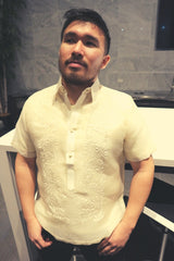 Lakhi stands in his hand embroidered cocoon Barong Tagalog with his black jeans. he stands in front of a white table and chairs with a marble wall in the background