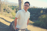Marc stands in his short sleeve hand embroidered cocoon Barong Tagalog. He also wears grey pants, glasses and a wrist watch. He stands in front of a tree on a hill. A ravine, trees, another hill and a view of the city of Los Angeles is in the background. Marc has his hands in his pockets and looks into the camera