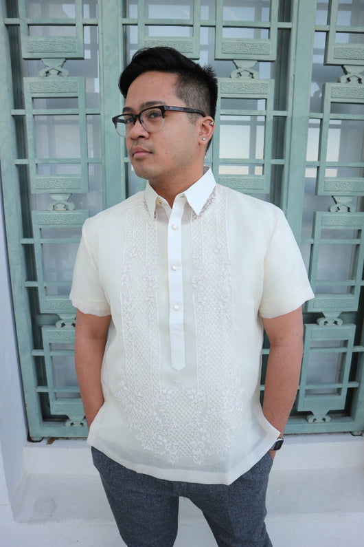 Marc standing in his short sleeve hand embroidered cocoon Barong Tagalog. He also wears grey pants, glasses and a wrist watch. He stands in front of a green decorative window at the Griffith Observatory. He has his hands in his pockets and his head is angled to his right