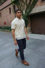 Matt walks on the sidewalk. He wears a short sleeve hand embroidered jusi Barong Tagalog. Matt also wears a chamisa de chino underneath his barong, navy blue pants, a watch on his left wrist, striped socks and light brown shoes. There is a brick garage covered in ivy in the background