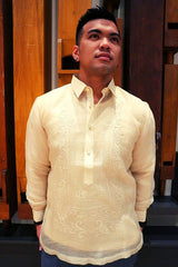 Michael stands in a hand embroidered cocoon Barong Tagalog, chamisa de chino underneath his barong, and grey pants. He stands with his hand in his back pockets looking up and to his left in front of wooden organ pipes.