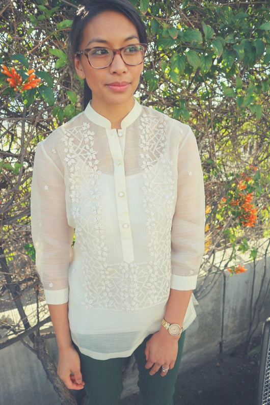Michelle stands in front of green leaves, brown vines, a few orange flowers and a concrete divider. She wears a hand embroidered piña silk Barong Tagalog, a white tank top underneath the barong, green pants, brown glasses and a watch and ring on her left wrist and hand..