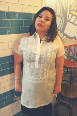 Natalia stands next to a dark green and white tiled pillar and in front of wooden stools, a wooden table and mural in the back. Natalia wears a dress length hand embroidered cocoon Barong Tagalog, a black tank top underneath the barong and black leggings.