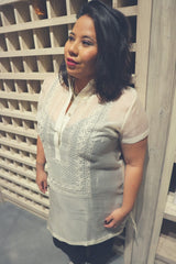 Angled photo of Natalia standing in front of an empty wine storage wall. Natalia wears a dress length hand embroidered cocoon Barong Tagalog. Natalia wears a black tank top underneath her barong and black leggings.  