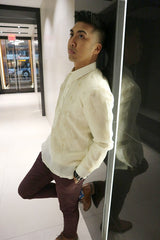 Noel stands leaning his back and right foot against a backlit black wall panel. There is a white hallway and glass door in the background. Noel wears a hand embroidered jusi Barong Tagalog, dark maroon slim slacks, blue patterned socks, brown dress shoes, a black watch on his left wrist