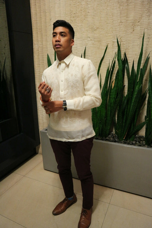 Angled photo of Noel standing in front of tall green plants in a rectangular long grey planter holder. There is beige textured wall behind him and a black wall and mirror to his right. Noel wears a hand embroidered jusi Barong Tagalog, dark maroon slim slacks, blue patterned socks, brown dress shoes, a black wrist watch on his left arm, and a Doom mask ring on his right hand. Noel adjusts his red and yellow bracelets on his right wrist with his left hand in front of him