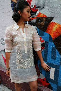Rachelle stands in her dress length hand embroidered piña silk Barong Tagalog in front of an Audrey Hepburn mural in Little Italy, New York City. She wears a beige tank top and black skirt underneath her barong. She stands at an angle in front of the wall with her left hand touching it. 