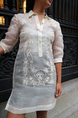 Product photo of the dress length hand embroidered piña silk Rachelle Barong Tagalog. Rachelle wears a beige tank top and black skirt underneath her barong. She stands in front of decorative wrought iron doors in New York City