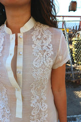 Closeup of the upper left side of the dress length hand embroidered piña silk Rechelle Barong Tagalog. She wears a beige slip dress underneath her barong. There is a gate, stacked rolls of wiring, steel bars and a suspended container in the background