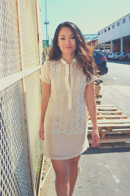 Rechelle walks towards the camera in her dress length hand embroidered piña silk Barong Tagalog. She wears a beige slip dress underneath her barong. She has a white gate to her right, pallets on the ground behind her. There are parked cars, buildings, a bridge and a tower in the background.