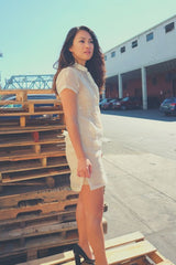 Side view of Rechelle standing on wooden pallets. Rechelle wears a dress length hand embroidered piña silk Barong Tagalog. She wears a beige slip dress underneath her barong. She also wears black high heel shoes. There is a pile of pallets behind Rechelle. There are parked cars, buildings, a bridge and a tower in the background.