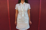 Product photo of the dress length hand embroidered piña silk Rechelle Barong Tagalog. Rechelle wears a beige slip dress underneath her barong. There is a perforated red wall behind her